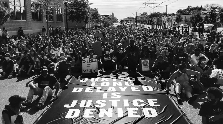 A crowd of people peacefully protesting in a street. A sign laying on the ground says "Justice Long Delayed is Justice Denied."